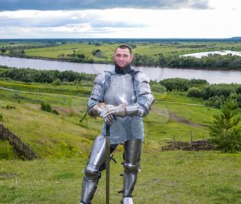 knight in armor stands against the background of native open spaces, forests and a river. Knightly armor and weapon. Semi - antique photo.