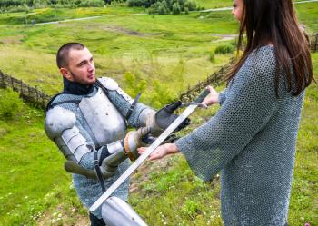 A lady in a chain mail hands a sword to her knight in armor. Knightly armor and weapon. Semi - antique photo.