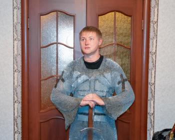 A man dressed up in a knight's hauberk at home in his room. Knight's sword in the hands of a man. Knightly chain armor and sword. Fitting of a knightly armor.