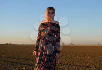 Woman in a plowed field in a red-black dress on a sunset background