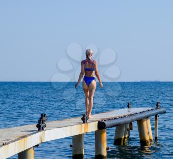 A beautiful girl in a blue bikini walks the pier to the sea. Marine concrete pier. Jumping into the water from the pier. Beautiful Booty Girls.