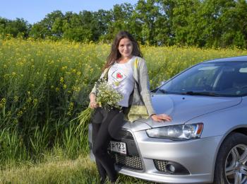 A beautiful young woman with a bouquet of daisies stands near a silver car.