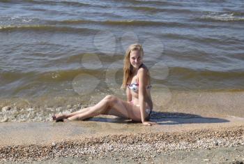 The girl the blonde in a bikini sitting on the beach in the sand. Beautiful young woman in a colorful bikini on sea background.