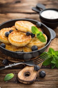 Delicious cottage cheese pancakes or syrniki with fresh blueberry in cast-iron pan on dark wooden rustic background. Tasty breakfast.