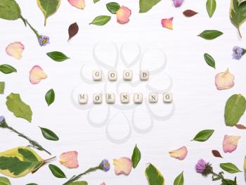 The phrase good morning on cubes in a frame of flowers, petals and green leaves on a white background.Inspirational image.Type flat, top view