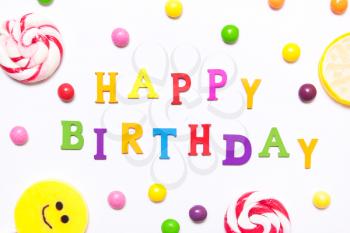 The phrase happy birthday, lollipops, candy smile on, are scattered around the colorful jelly beans on a white background. Top view, flat