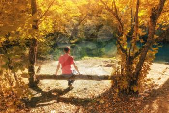 Man sits on the branch of tree near river at auntumn forest. Nature landscape scene.