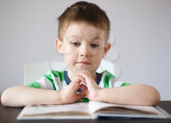Cute little boy is reading book while sitting at table