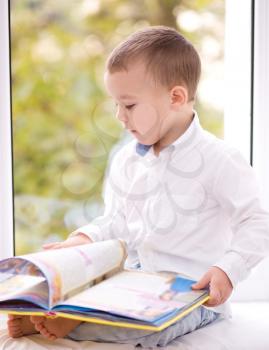 Little boy is reading book while sitting on windowsill, indoor shoot