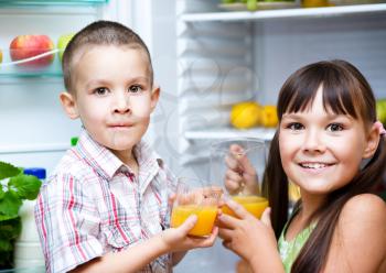 Happy children drink orange juice standing near refrigerator with fruits and vegetables