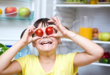 Happy girl eating tomatoes standing near refrigerator with fruits and vegetables