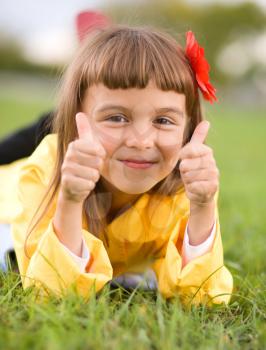 Little girl lying on green grass is showing thumb up gesture