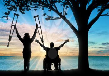 Happy people with disabilities in wheelchair and on crutches on beach standing under tree. Concept happy disabled