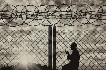 Concept of the religion of refugees. Silhouette refugee praying near the fence with barbed wire on the background of evening city