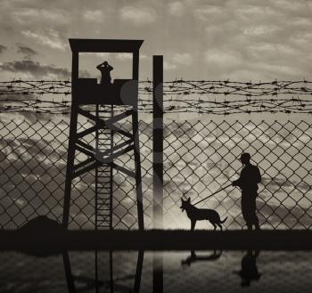 Concept of security. Silhouette of a lookout tower and a guard with a dog on the background of the fence with barbed wire and reflection