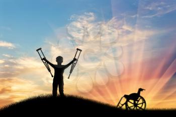 Happy child with crutches disabled person next to wheelchair on hill and sunset. Concept of disability