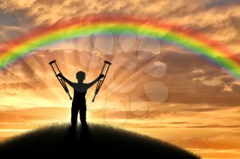 Child disabled with crutches on a hill on a background of a sunset rainbow. Concept of disability