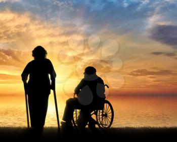 Silhouette of an old woman on crutches and elderly man in a wheelchair on a background of sea sunset. Concept of disability and old age