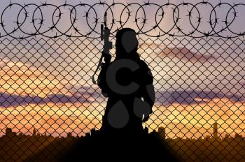 Concept of terrorism. Silhouette terrorist near the border fence in the background of the city at sunset