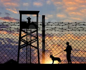 Concept of security. Silhouette of a lookout tower and a guard with a dog on the background of the fence with barbed wire at sunset