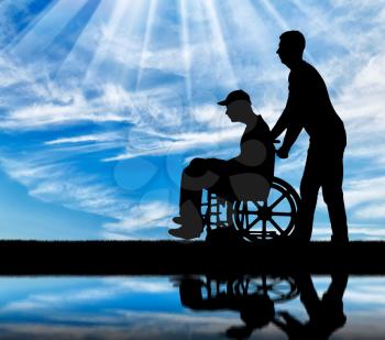 Silhouette of a walk of a disabled man in a wheelchair and a close person against the sky by the river. The concept of caring for people with disabilities