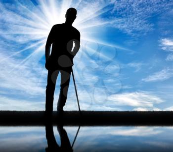 Silhouette of a disabled man supports himself with a crutch near the river with his reflection. Concept of people with disabilities