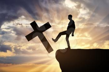 Man atheist pushes the symbol of christian cross into the cliff on sunset background. Concept of atheism