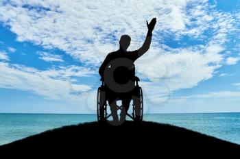 Silhouette of a disabled man in a wheelchair friendly waving his hand atop a hill near the sea. The concept of people with disabilities
