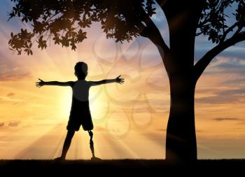 Children with disabilities concept. Happy disabled boy with a prosthetic leg standing near a tree at sunset