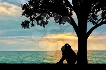 Silhouette of sad little girl sitting under a tree by the sea crying. Conceptual image of parents' divorce