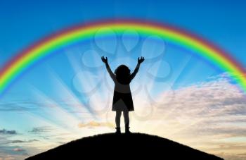 Silhouette of a happy little girl child with raised arms trying to touch the rainbow. A conceptual scene of a happy childhood