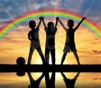 Childhood concept. Three happy children holding hands on a rainbow background and the sunset over the water with their reflection