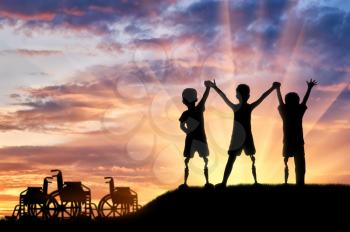 Children's disability concept. Happy children with disabilities with a prosthetic leg standing, holding hands at sunset, and wheelchairs
