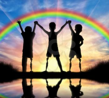 Children's disability concept. Children with disabilities with a prosthetic leg standing, holding hands on a background of a rainbow at sunset near the river