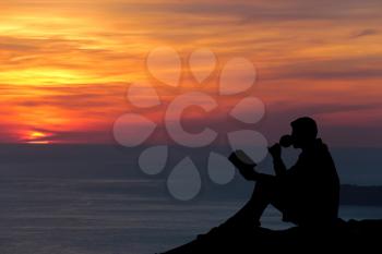Silhouette of a man sitting on breakwater in evening near sea, reads book  and drink wine
