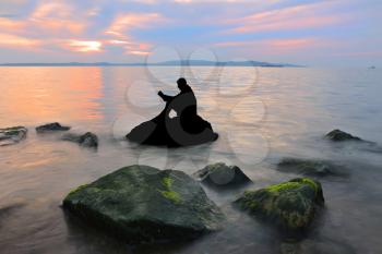 Silhouette guy sitting on breakwater in evening near sea, reads book