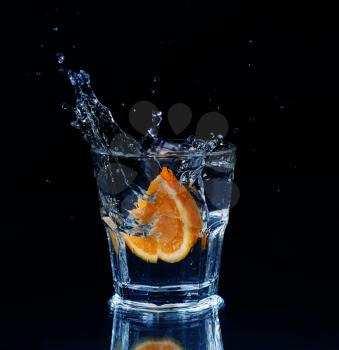 Slice of lemon splashing into a glass of water with a spray of water droplets in motion suspended in the air above the glass on a dark background.