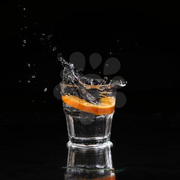 Half of orange falling down in glass with water on deep black. Beautiful glass with sparkling water or other transparent drink and a slice of lemon on black background