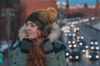 Beautiful winter portrait of young woman in the winter snowy scenery. Pretty red-haired girl walking on a winter evening city