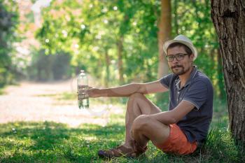 Smiling man sitting in forest looking at camera. guy with water. near the tree and backpack