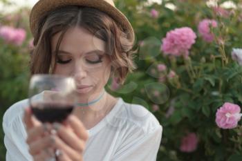 Beautiful young woman with curly hair posing near roses in a garden. Girl with a glass in her hand, tasting red wine and having fun
