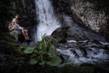 Young barefooted tourist wearing jeans short and backpack sitting on big rock looking peaceful and relaxed with waterfall on background