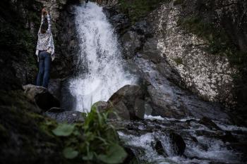 Boho woman wearing hat and poncho standing by the waterfall and looking at it. Cold weather, fall hiking. Wanderlust photo series.