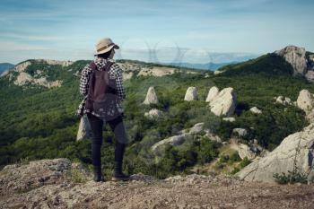 woman traveler with backpack holding hat and looking at amazing mountains and forest, wanderlust travel concept, space for text, atmospheric epic moment