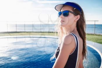 beautiful girl with perfect tanned body in in blue one piece bathing suit and hat, sunlight sunglasses relaxing beside a swimming pool