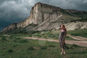 attractive young woman outdoors with stone background. Red-haired girl in ethnic dress on a background of a stormy sky. Belogorsk, White Rock