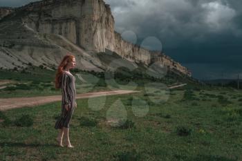attractive young woman outdoors with stone background. Red-haired girl in ethnic dress on a background of a stormy sky. Belogorsk, White Rock