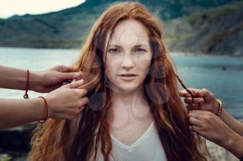 Red-haired girl with hippies with freckles, whose hair is braided by two friendsCrimea, Fox bay, Koktebel