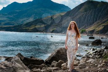 Beautiful boho styled model wearing white dress posing on the beach in sunlight. Red-haired girl with freckles. Crimea, Fox bay, Koktebel