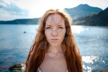 Beautiful boho styled model wearing white dress posing on the beach in sunlight. Red-haired girl with freckles. Crimea, Fox bay, Koktebel